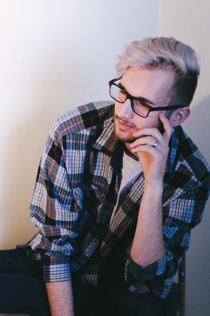 man wearing black, blue, and red plaid collared button-up long-sleeved shirt and eyeglasses sitting and putting left hand on his face